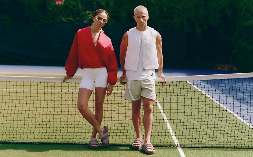 A man and a woman standing on opposite sides of a tennis net on a green tennis court. The woman wears a bright red long-sleeve top, white shorts, and sandals, while the man wears a white sleeveless shirt, light beige shorts, and sandals.