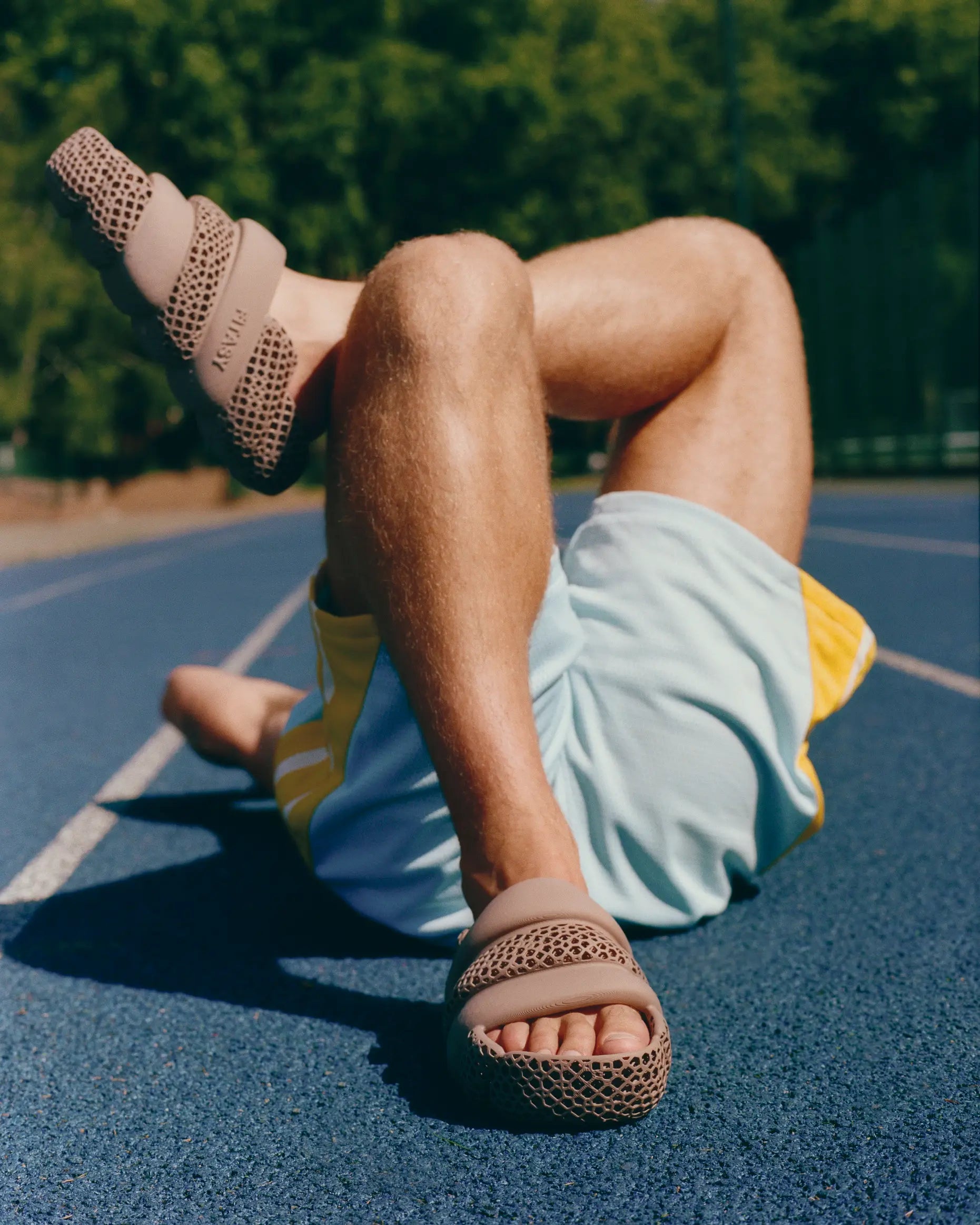 A person lying on a blue athletic track field wearing beige woven slide sandals and light blue shorts with yellow side panels. Trees are visible in the background.