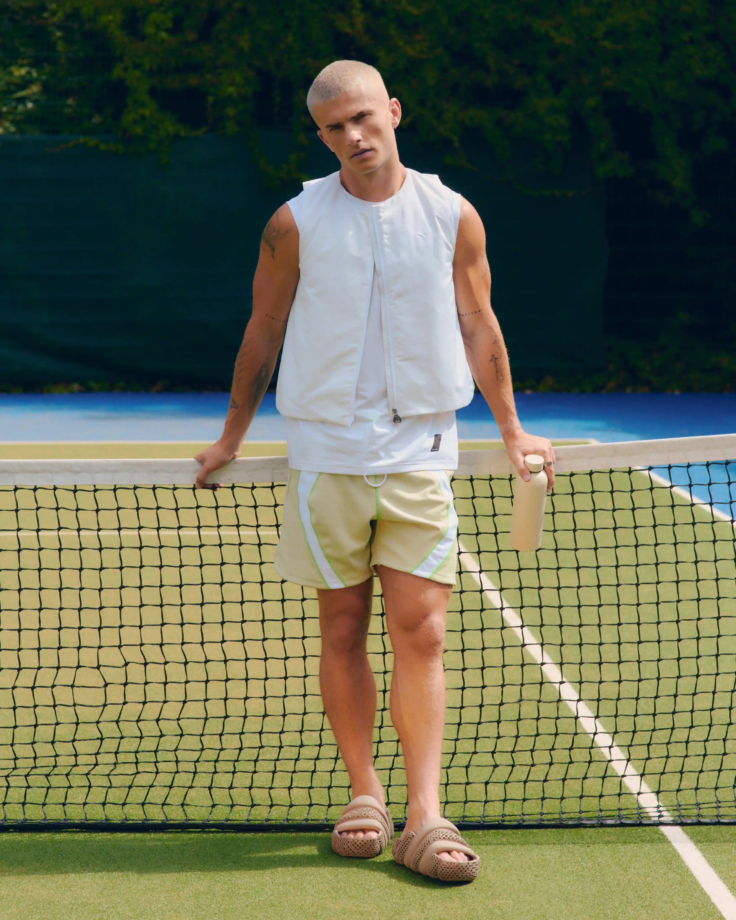 A man standing on a tennis court behind the net, wearing a white sleeveless zip-front top, light beige shorts, and tan sandals.