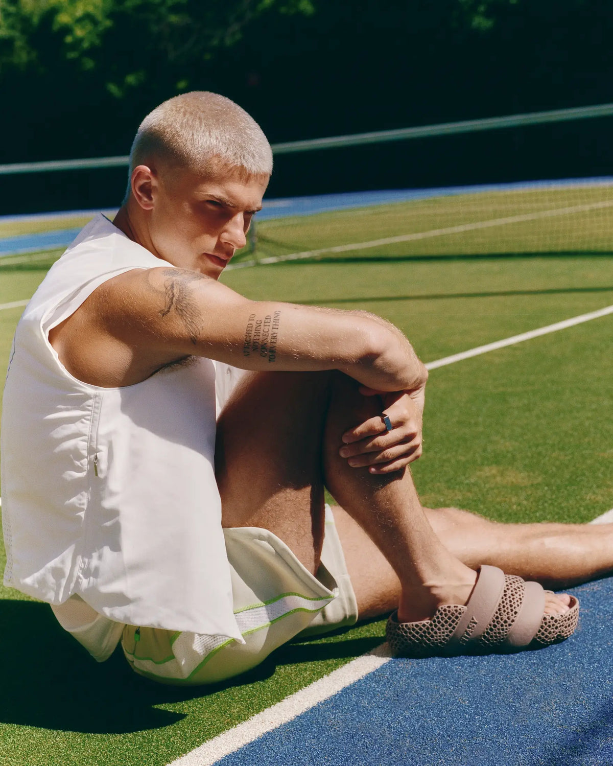 A man with a buzz cut sits on a blue tennis court wearing a white sleeveless shirt, light green shorts, and beige perforated slide sandals.