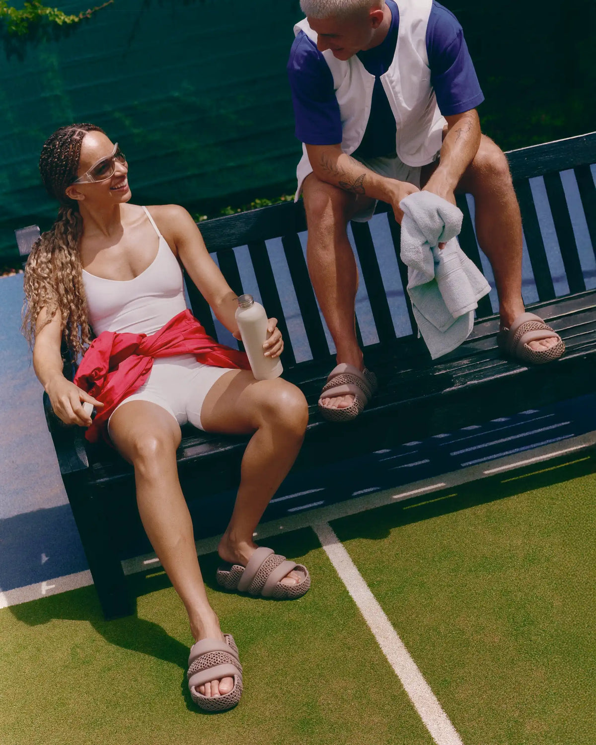 A woman and a man sitting on a black slatted bench on a tennis court. The woman wears a white tank top, white shorts, and a red shirt tied around her waist, while the man wears a white and navy shirt and shorts.