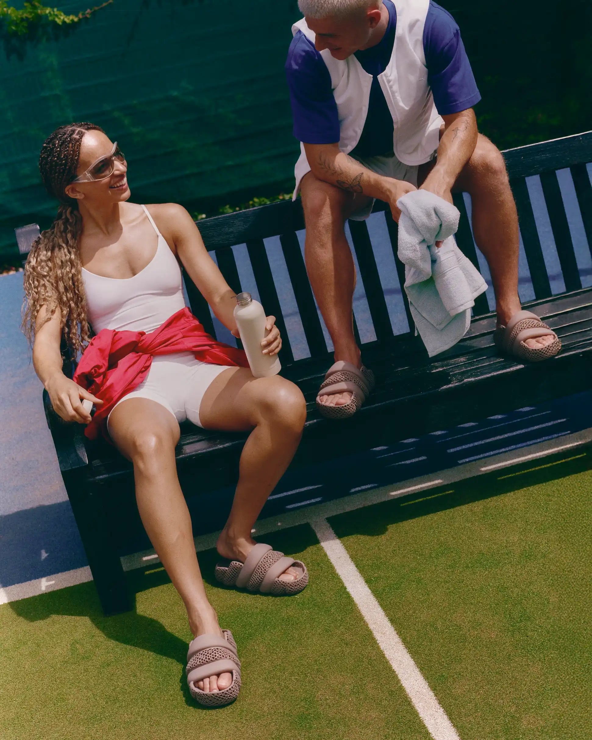 A woman and a man sitting on a black slatted bench on a tennis court. The woman wears a white tank top, white shorts, and a red shirt tied around her waist, while the man wears a white and navy shirt and shorts.