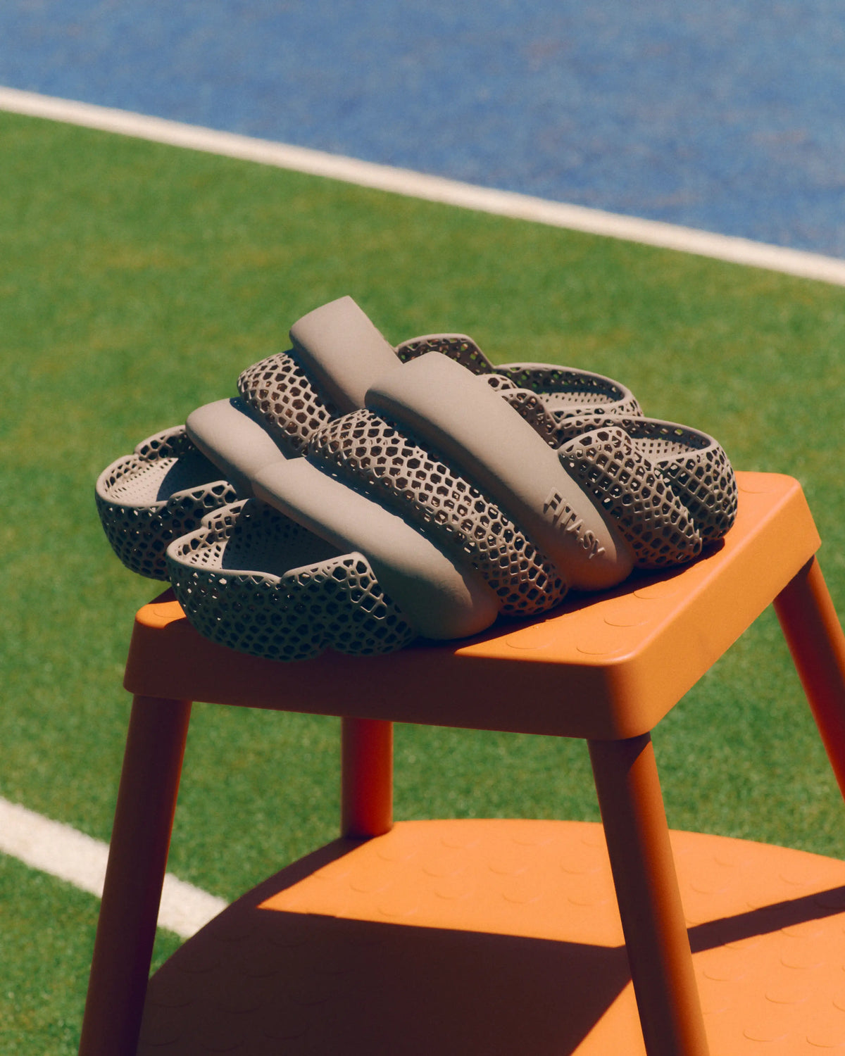 A pair of chunky, sculptural slide sandals with a beige smooth upper and a dark gray honeycomb-patterned mesh lower band, displayed on an orange stool on an outdoor sports field.