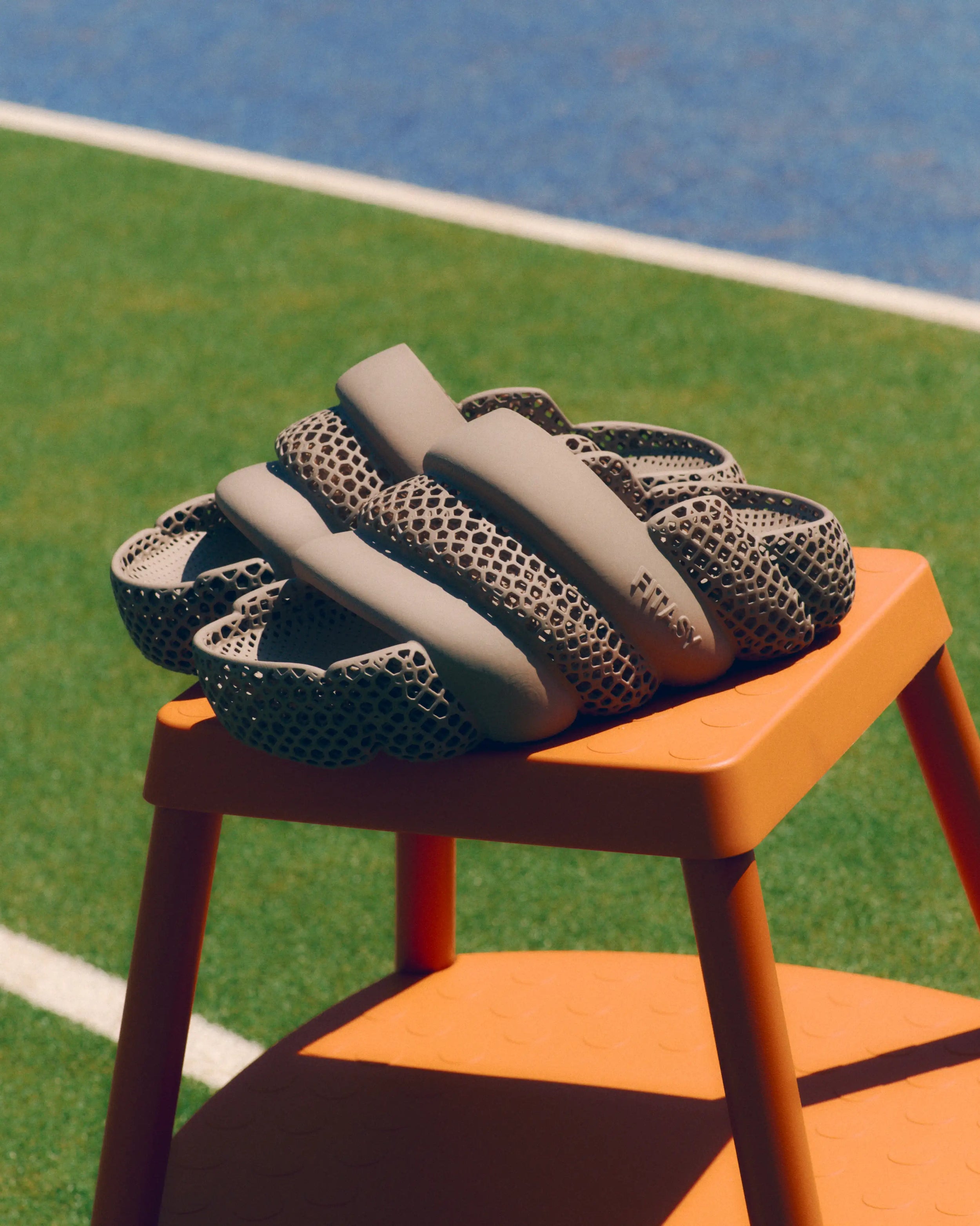 A pair of chunky, sculptural slide sandals with a beige smooth upper and a dark gray honeycomb-patterned mesh lower band, displayed on an orange stool on an outdoor sports field.